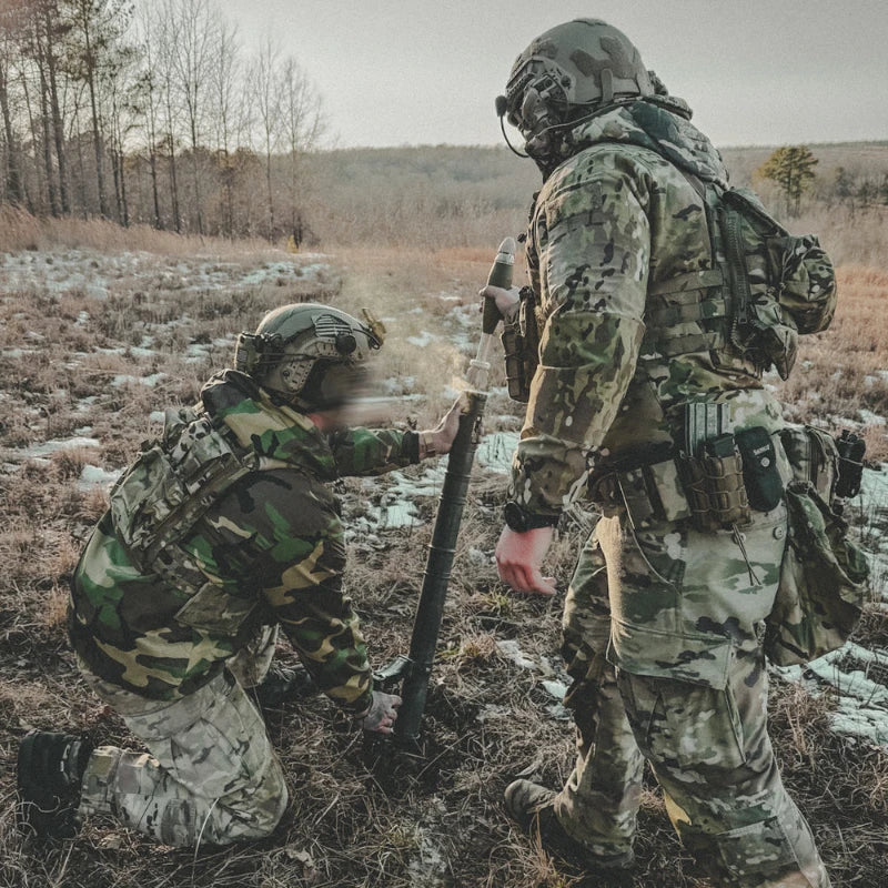 Two soldiers in camouflage gear working together in a field.
