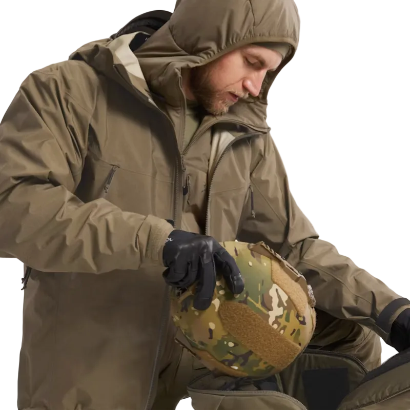 A man wearing a hooded olive green tactical jacket and black gloves holds a camouflage combat helmet with a tan velcro patch, against a plain light background.