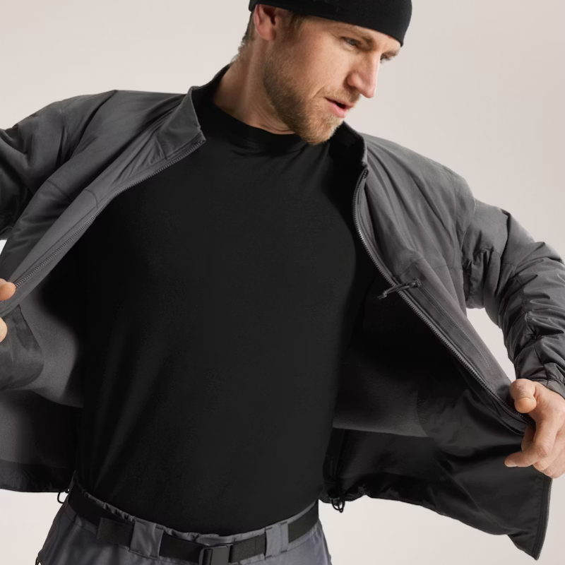 A man wearing a black crewneck base layer shirt and a dark gray zip-up jacket, opening the jacket to show the interior. He is also wearing a black beanie and gray pants against a plain light gray studio background.