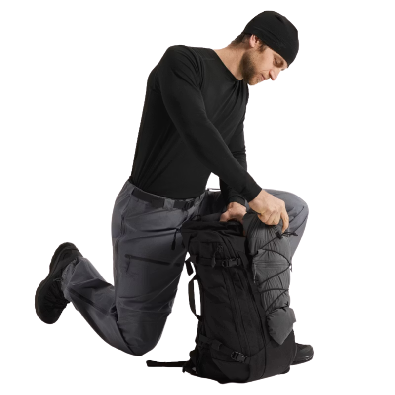 A man in a black beanie, black long-sleeve shirt, and gray pants kneels beside a black technical backpack, opening the top zipper area. The background is solid white.