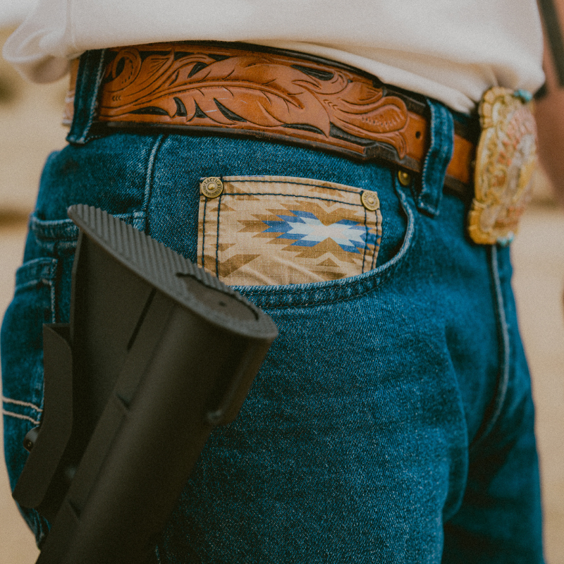 Person wearing blue jeans with a decorative pocket, brown leather belt, and holding a black rifle.
