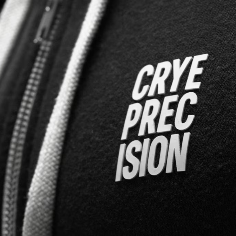 Close-up of a black zippered garment with a white zipper pull and a white embroidered patch reading 'CRYE PRECISION'.