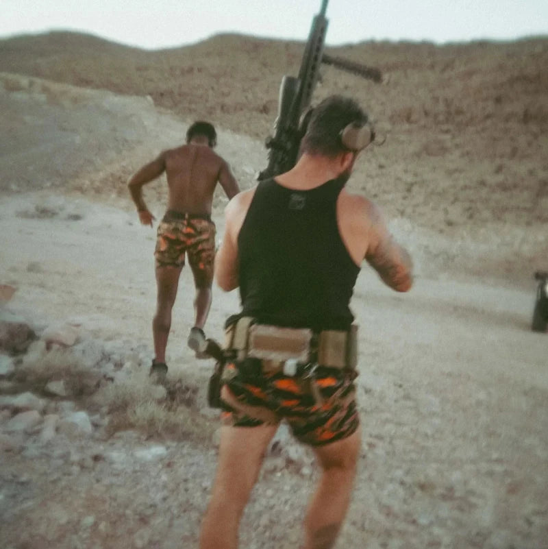 Two men in a desert setting with one holding a rifle. Both are wearing blaze orange tiger stripe shorts