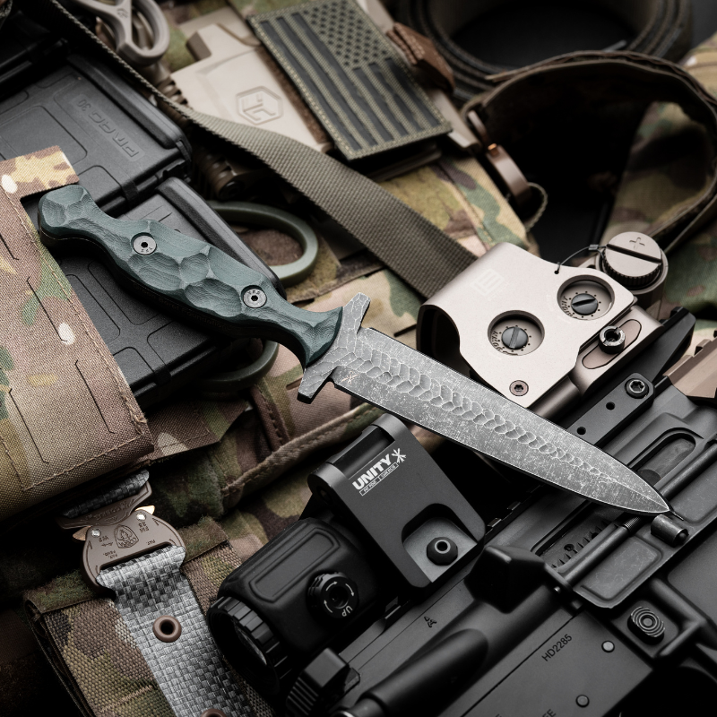 Tactical fixed-blade knife with acid-dipped double-edge blade and sculpted G10 handle displayed on a camo plate carrier alongside rifle optics, magazines, and gear — showcasing elite military-grade EDC equipment for field-ready performance.