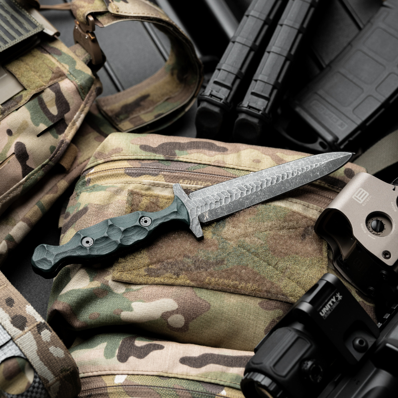 Tactical fixed-blade knife with acid-dipped double-edge blade and sculpted G10 handle displayed on a camo plate carrier alongside rifle optics, magazines, and gear — showcasing elite military-grade EDC equipment for field-ready performance.