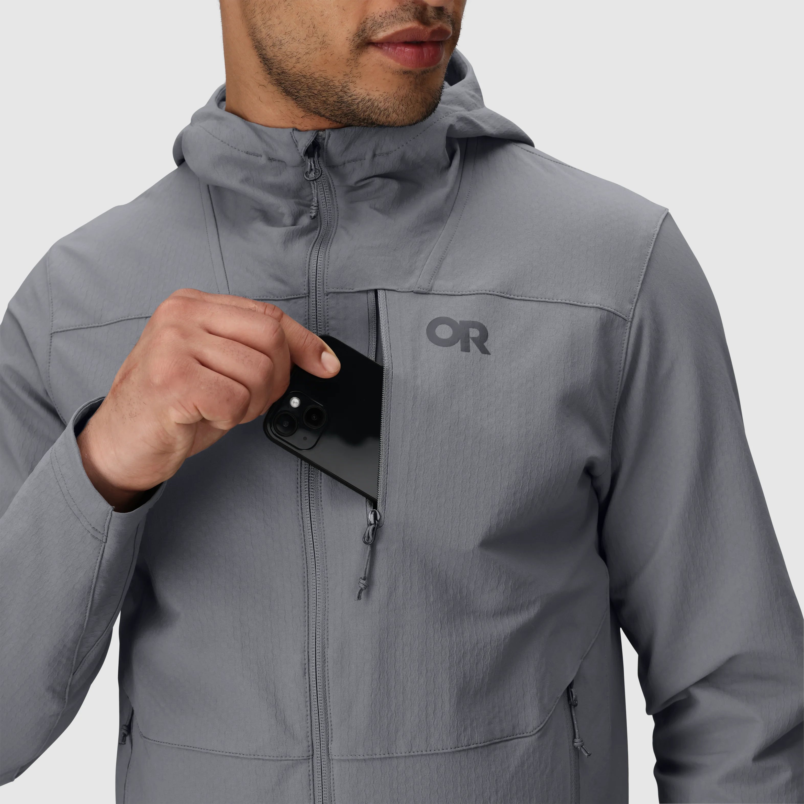 Detail view of the Outdoor Research Ultima Soft Shell Hoodie showing a phone being placed into the zippered chest pocket, highlighting secure storage and durable weather-resistant zippers for alpine and trail use.