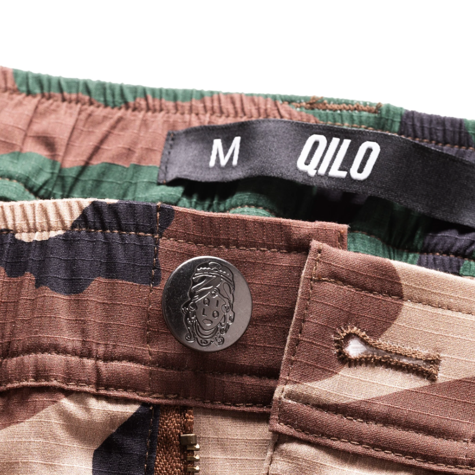 Detail view of QILO x WRMFZY collaboration shorts showcasing custom logo button and tactical upgrades. Woodland Camo Shorts Limited Edition.