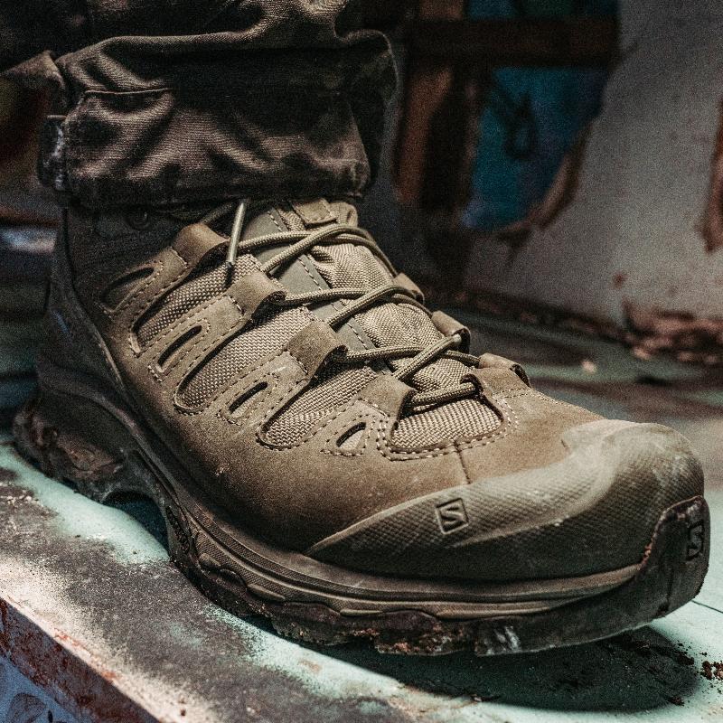On body view of the Salomon FORCES Quest 4D 2 High GTX boots being worn in muddy rugged terrain showing the waterproof GORE TEX construction, aggressive outsole traction, and high support design built for military operations, extended rucking, and harsh field environments.