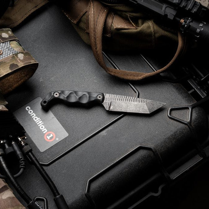 Lifestyle flat lay of the Stroup Knives Bravo 5 Tanto with acid-etched blade, showing the full-tang 1095 steel construction, rugged fixed-blade profile, and field-ready design from the Stroup Knives x Justin Melnick collaboration, arranged with tactical everyday carry gear.