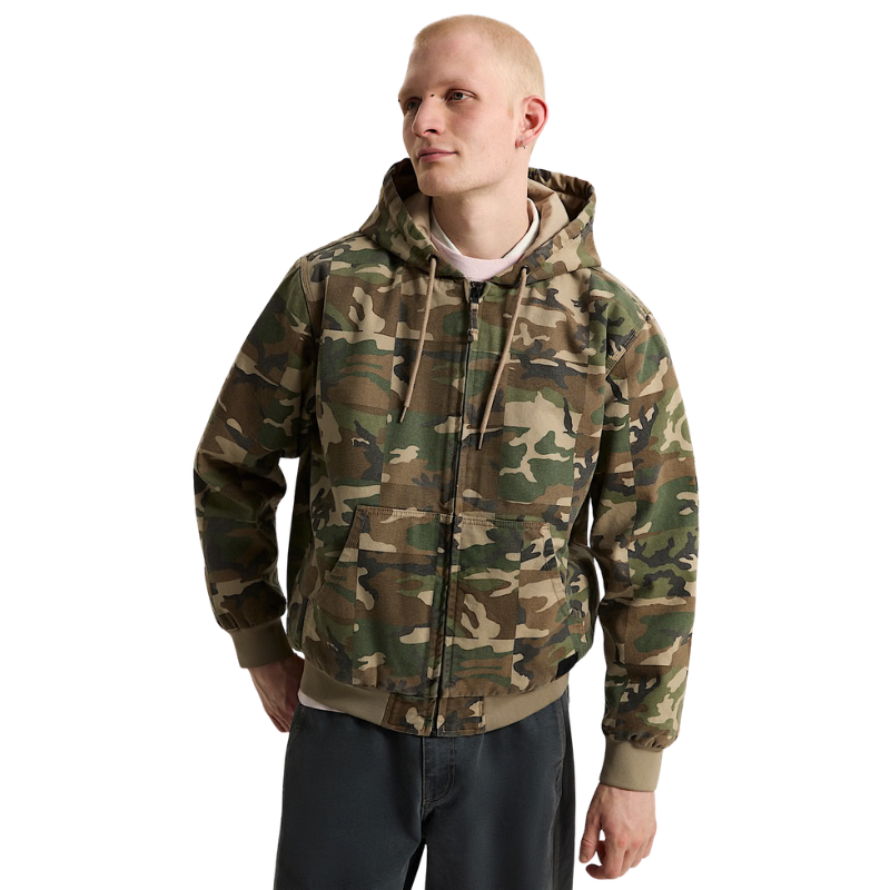 A man wearing a camouflage zip-up hooded jacket over a white shirt and dark pants, photographed against a plain white studio background.