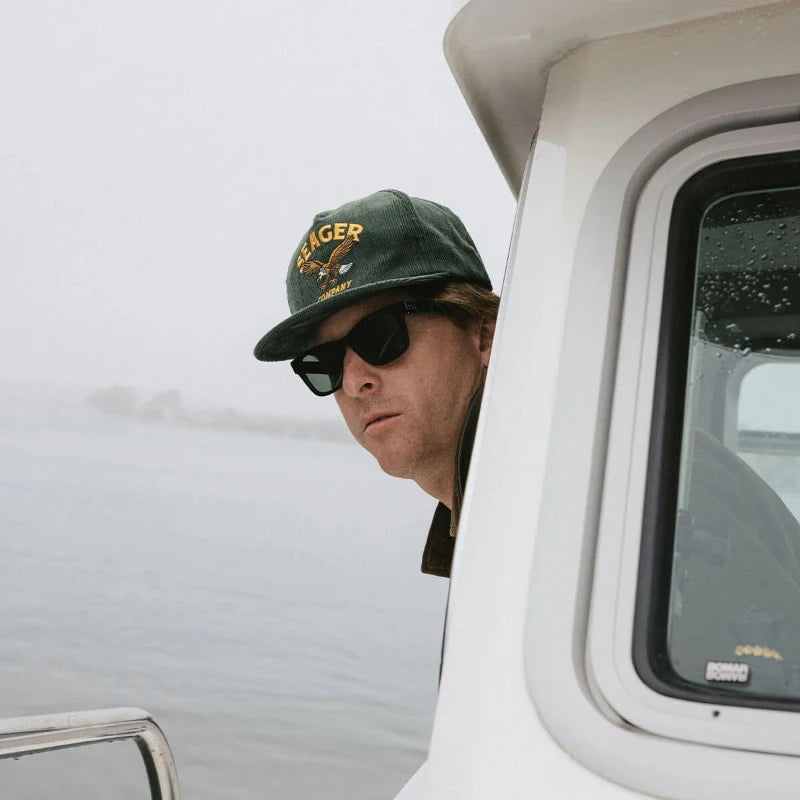 Person wearing a cap and sunglasses, peeking out of a boat window with a blurred background