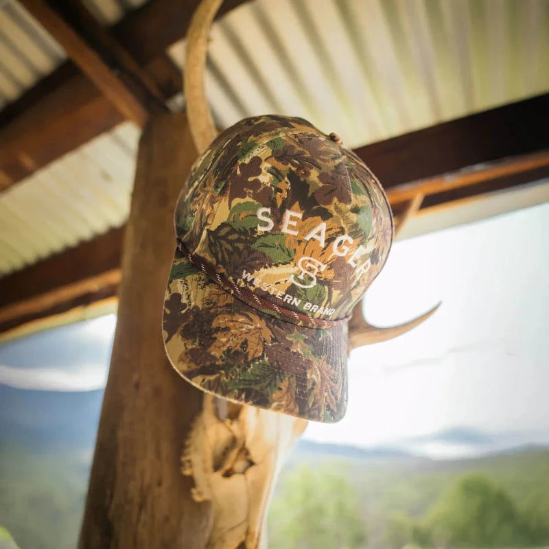 Camouflage cap with 'SEAGER' logo hanging on taxidermy. Hung on a wooden surface with a scenic background