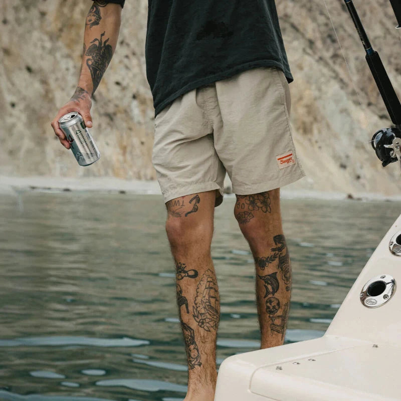 Person with tattoos holding a can by water, wearing beige shorts
