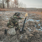 Person in camouflage gear using a tool in a snowy field with trees in the background