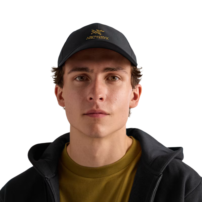 A young man wearing a black Arc'teryx baseball cap, a black zip-up hoodie, and a mustard yellow t-shirt, photographed against a solid white background.