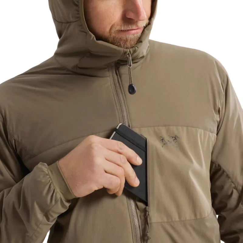 A close-up of a man wearing a hooded Arc'teryx jacket in a warm khaki-tan color, demonstrating a zippered chest pocket that holds a black cardholder.