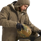 A man wearing a hooded olive green tactical jacket and black gloves holds a camouflage combat helmet with a tan velcro patch, against a plain light background.