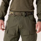 Close-up of a person wearing olive green tactical cargo pants with a camouflage-patterned belt and olive green shirt, shown against a light neutral background.