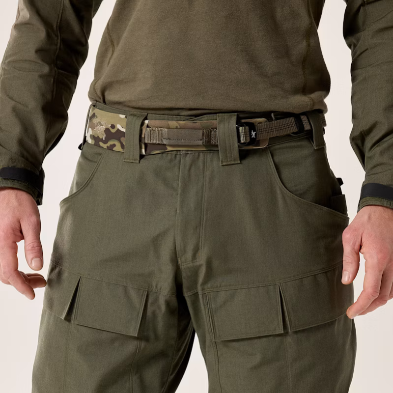 Close-up of a person wearing olive green tactical cargo pants with a camouflage-patterned belt and olive green shirt, shown against a light neutral background.