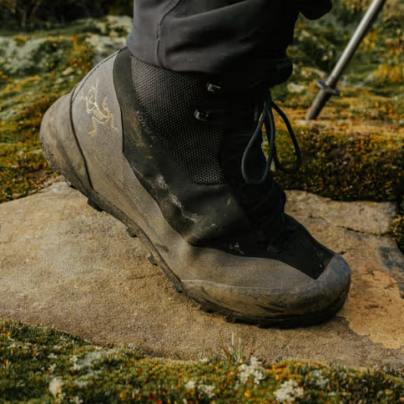 A close-up of a person's foot wearing a black Arc'teryx hiking boot with a muddy gray sole, standing on a mossy stone outdoors.