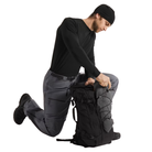 A man in a black beanie, black long-sleeve shirt, and gray pants kneels beside a black technical backpack, opening the top zipper area. The background is solid white.