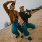 Two men in outdoor attire with one holding a rifle on a sandy hill.