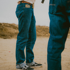 Two people wearing blue jeans standing on a sandy surface with a blurred background