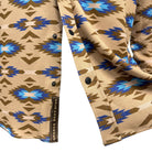 Close-up of a patterned button-up shirt with a Southwestern geometric design in beige, brown, blue, and light blue. A black button is visible, and a vertical fabric label reads 'HOLD YOUR HORSES'.