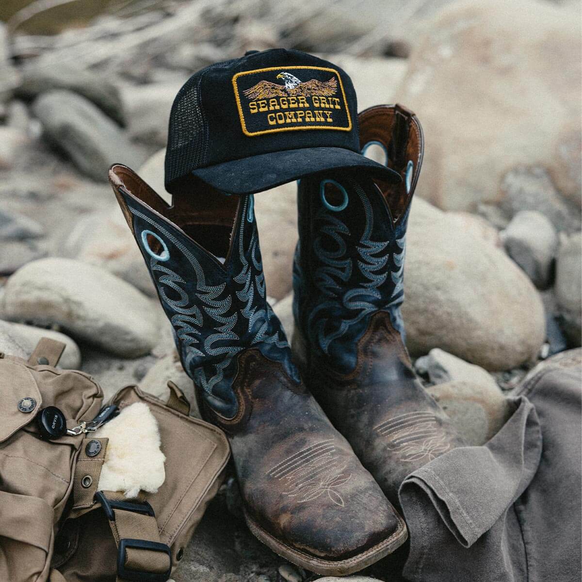 Pair of cowboy boots and a Seager cap on rocks with outdoor gear.