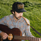 Man playing guitar outdoors in a grassy field wearing a cap with a Seager logo.