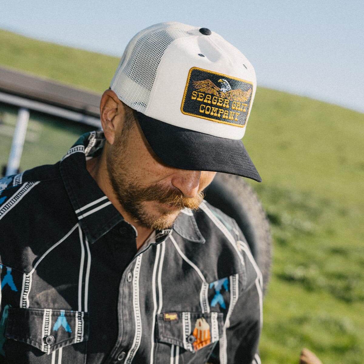 Man wearing a cap with a Seager logo, standing outdoors with green grass and blue sky in the background.