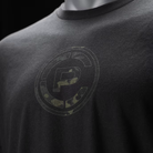 Close-up of a black t-shirt on a mannequin, featuring a distressed circular graphic with a stylized letter 'CP' in Multicam Black.