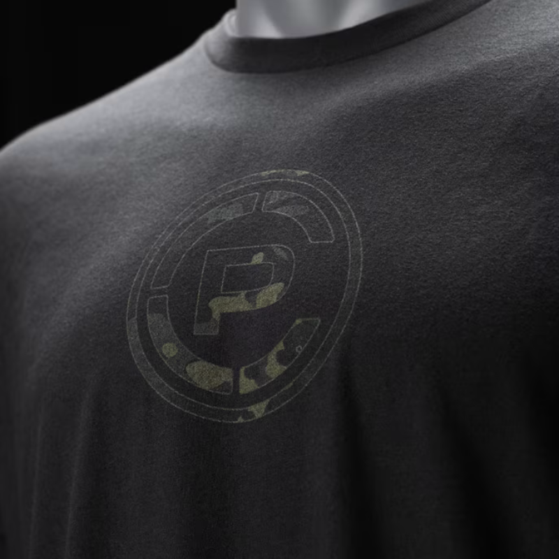 Close-up of a black t-shirt on a mannequin, featuring a distressed circular graphic with a stylized letter 'CP' in Multicam Black.