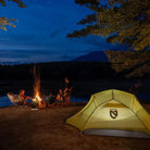 Camping scene with a yellow tent, campfire, and people around a lake at night.