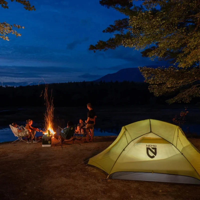 Camping scene with a yellow tent, campfire, and people around a lake at night.