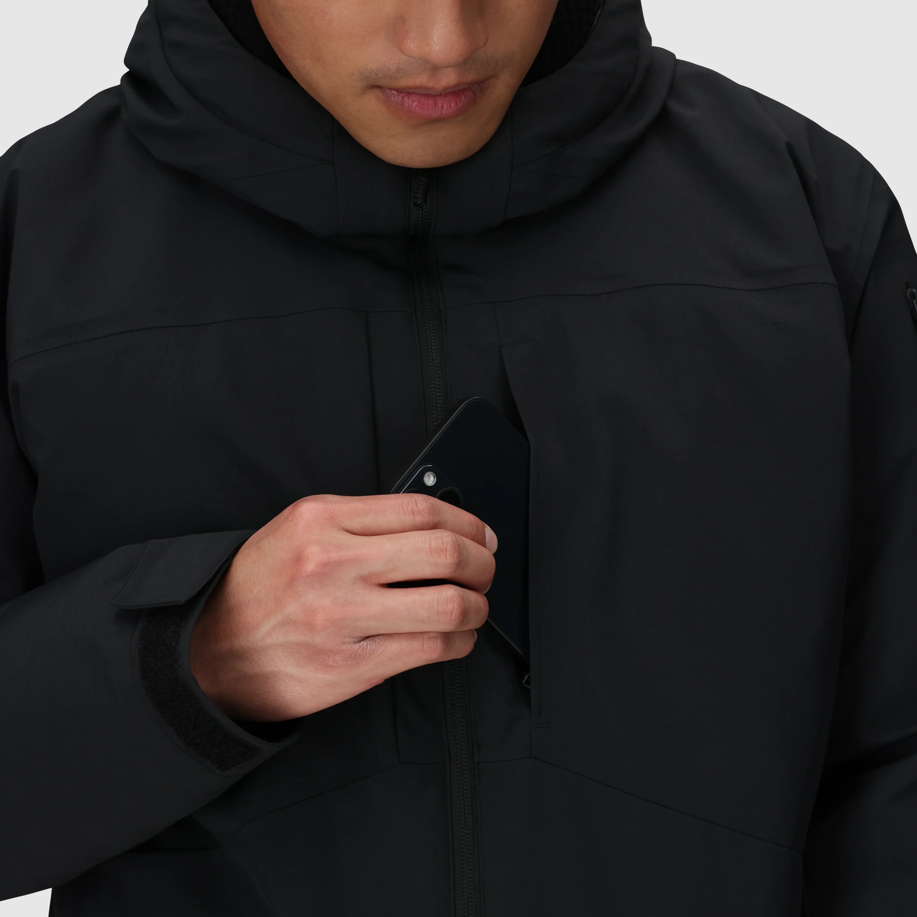 Close-up of the Outdoor Research Allies Colossus II Parka showing a phone being placed into the front chest pocket, highlighting secure waterproof storage designed for tactical gear, survival essentials, and cold-weather operations in Arctic conditions.