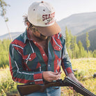 Man holding a rifle in a field with mountains in the background, wearing a 'Seager' cap.