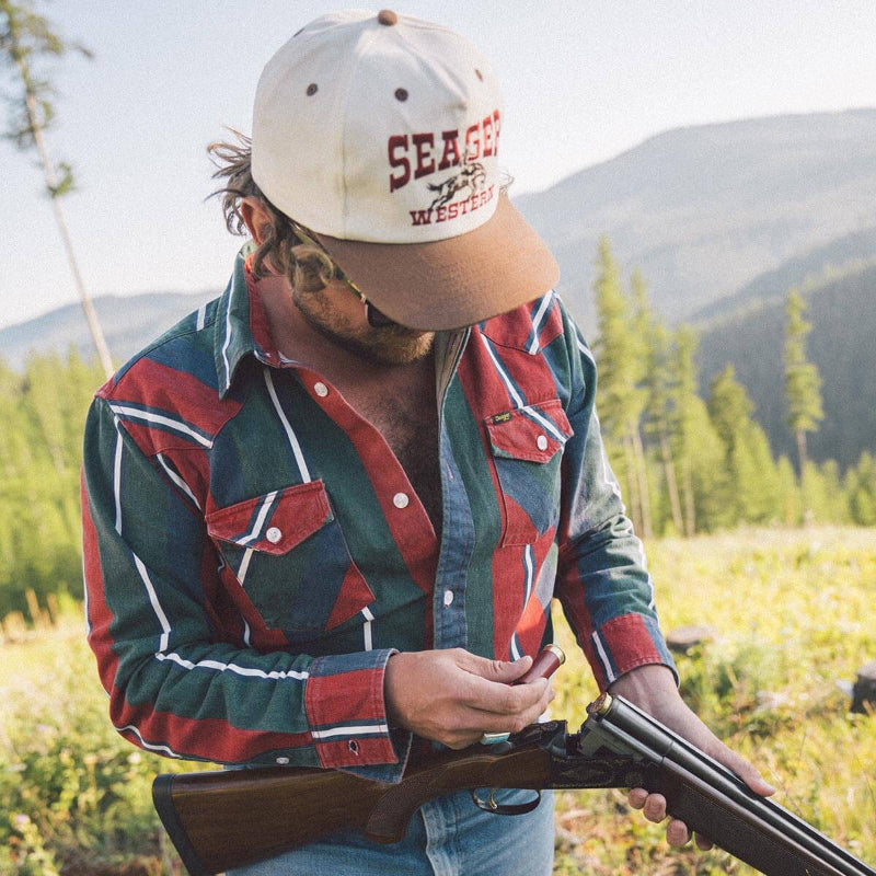Man holding a rifle in a field with mountains in the background, wearing a 'Seager' cap.