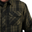 Close-up of a man wearing a plaid button-up shirt in dark olive green and black, featuring a chest pocket with a flap and a visible Tac-Dis brand label.