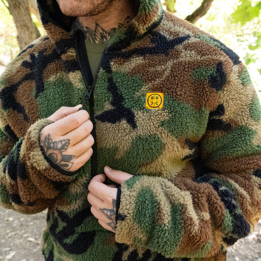 Close-up detail of the TD x Black Rifle Coffee Co. Reticle Half-Zip Fleece highlighting the woodland camo Sherpa fleece texture, half-zip front, and co-branded TD x BRCC logo patch. Durable birdseye mesh trim adds structure and breathability to this premium tactical fleece built for warmth, comfort, and all-day performance.