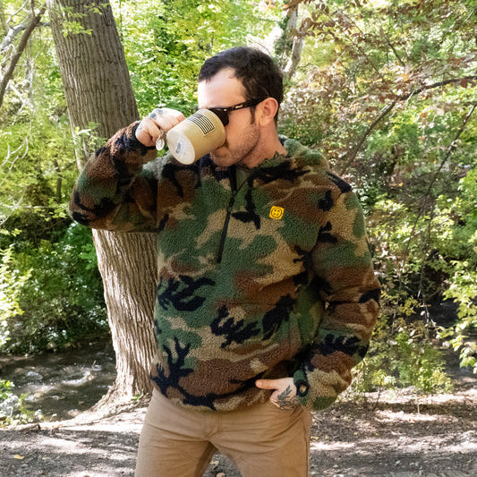 Man wearing the TD x Black Rifle Coffee Co. Reticle Half-Zip Fleece in woodland camo while drinking coffee, showing off-duty comfort meets tactical grit. High-pile Sherpa fleece with breathable mesh trim built for warmth, mobility, and caffeine-fueled mornings. Co-branded TD x BRCC design made for range days, campfires, and cold-weather missions.
