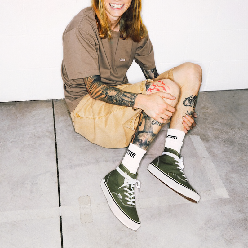 Person sitting on a concrete floor wearing a brown shirt, beige shorts, and green sneakers.