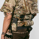 Person wearing a camouflage tactical vest with various pouches and equipment on a plain background